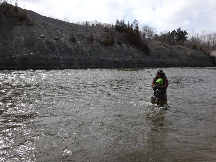 Two anglers wading a broad Southern Ontario river below a shale cliff, one with a rod bent into a fish